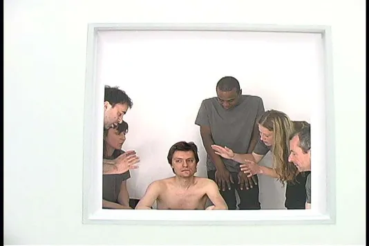 5 performers gathered around a shirtless man in a video still.