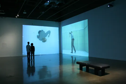 2 people stand in front of huge video projections in a corner. Both videos show a man in motion, one from a birds eye view.