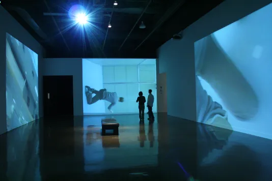 2 people stand shadowed in the blue hue of huge video projections covering the walls around them with reflections on the floor.