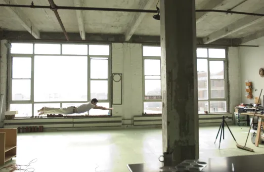 A man in white clothing floats midair with arms and legs extended in a bright open studio room with large windows.