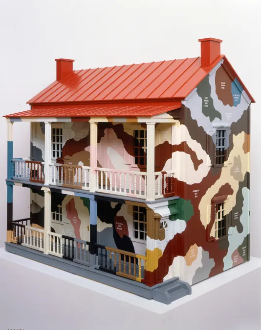 Close view of a model house with a bright red roof, and outer walls painted in a colorful camouflage pattern on a pedestal.