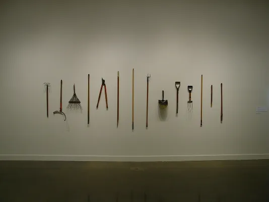 A variety of 14 garden tools hung at differing heights on a white wall, equally spaced, pointed up and down, casting shadows.