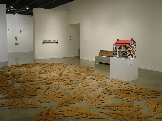 Leaf shaped stenciled cocoa mats grouped together and spread over gallery floor, next to house sculpture and other artworks.