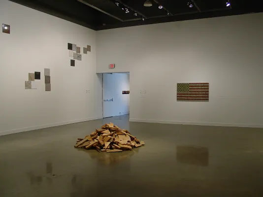 Scrap wood piled on the floor, groupings of color square stones and a sculpture resembling a U.S. flag hang on walls behind.
