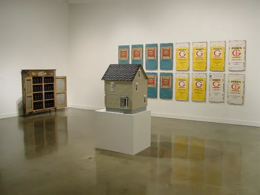 A drab colored model house on a pedestal, a cabinet with open doors to the left, and a group of bright artworks hang behind.