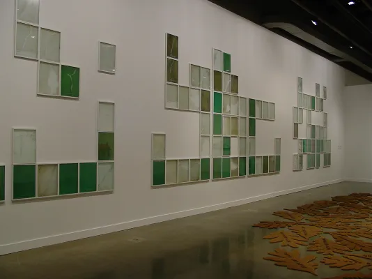 Large installation of 3 groupings of green hued glass panels arranged in abstract forms across a wall, with mats on floor.