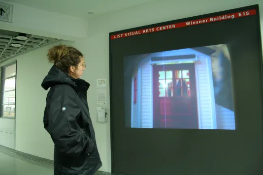 A visitor stands in front of a video projection in a hallway showing a blurry view of a red door with a person visible behind it.
