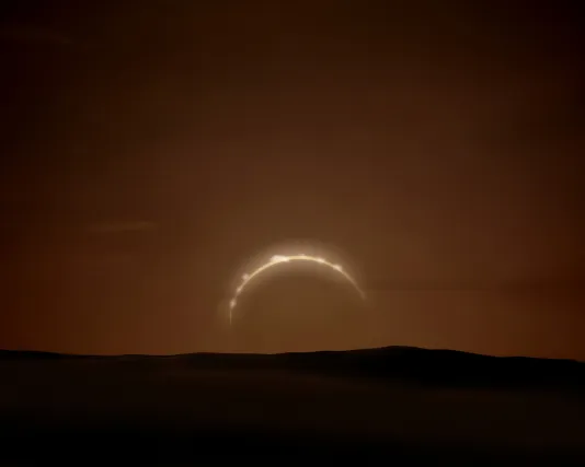 Amidst a total solar eclipse, a sliver of sunlight gives shape against a brown coloured sky with a dark shadowy landscape below.