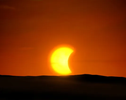 A bright yellow sun is partially eclipsed by the moon against an orange, red sky with a dark shadowed landscape directly below.