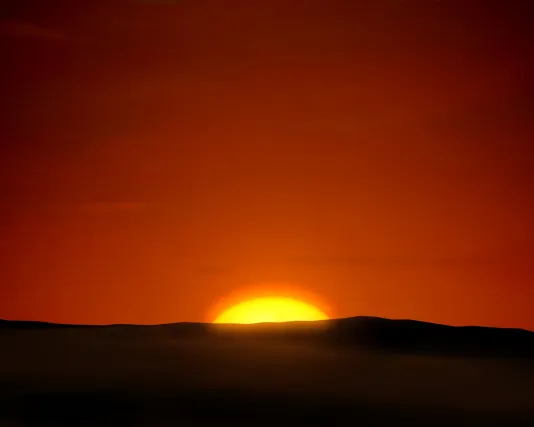A partially visible yellow sun glows bright against a vibrant orange, red sky with a dark shadowed landscape directly in front.