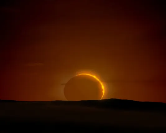 A sliver of yellow sunlight gives shape to a total solar eclipse against a brown coloured sky with a dark shadowy landscape below.