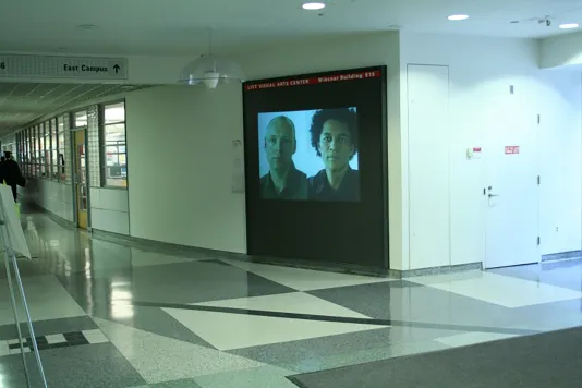 A view of a projection space in a hallway at MIT showing a portrait of the artist and man side by side.