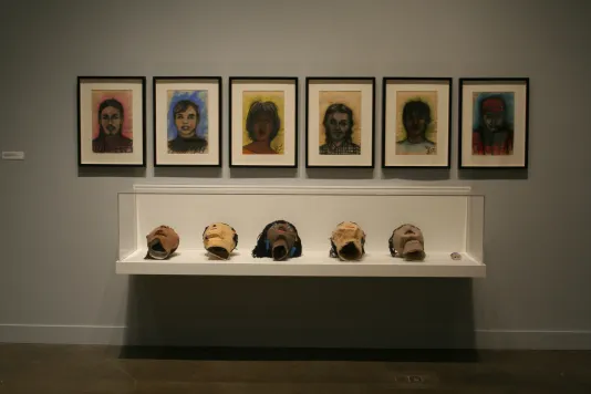 Six colorful portrait drawings hanging above a glass case containing 5 papier-mâché heads, laying flat, & a ceramic figurine.