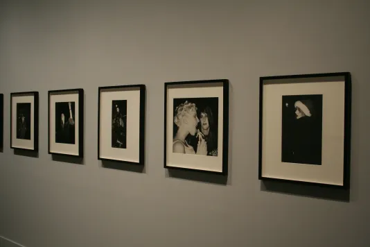 A series of black and white photographs hang on a wall. The most visible photo shows a woman holding and licking a small doll.