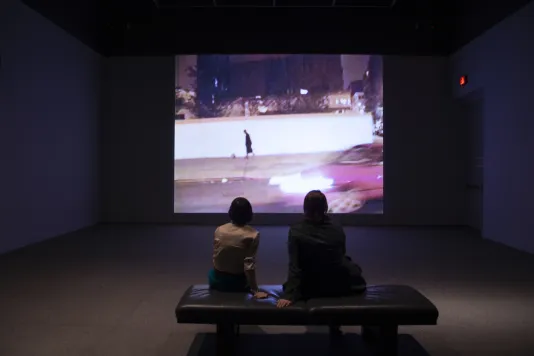 Two people sit on a gallery bench and watch a video projection with a moving red car and a person walking.