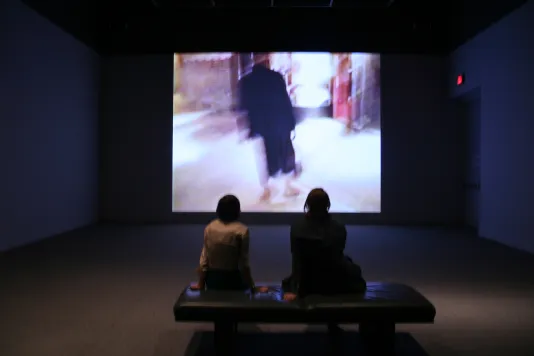 Two people sit on a gallery bench and watch a video projection with a blurry person in a suit.