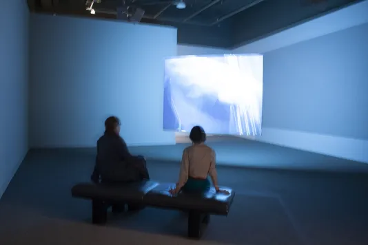 Two people sit on a gallery bench and watch a video projection with a bright and blurry blue light.
