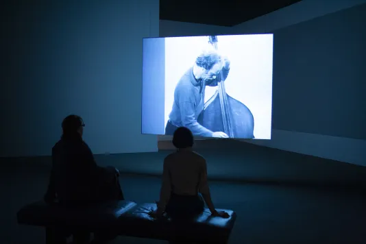Two people sit on a gallery bench and watch a video projection of a man playing the upright bass.