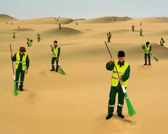 Identical men wearing green uniforms and Day-Glo™ work vests sweep sand in an infinite desert.