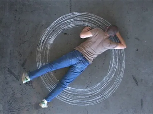 An aerial view of an artist stretching out on the floor with a piece of chalk. The artist draws concentric circles.