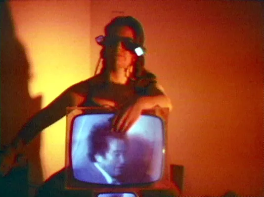 Red hued image of a woman sitting with a retro television monitor in her lap. She is wearing glasses and the monitor depicts a man in a suit looking down.