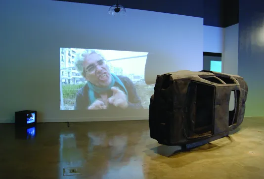 A black burnt out car on its side, a wall projection of a man pointing his finger and grimacing, a box video monitor on the floor