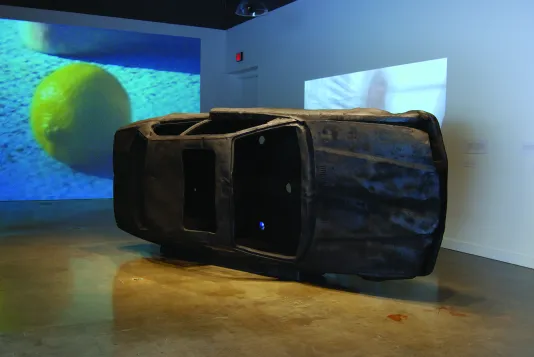 A black burnt out car on its side, and two video projections, one with an image of a lemon