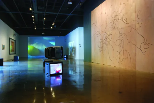 A darkened space with box monitors on the floor, a large wall drawing on wood, projections, and a burnt out car on its side