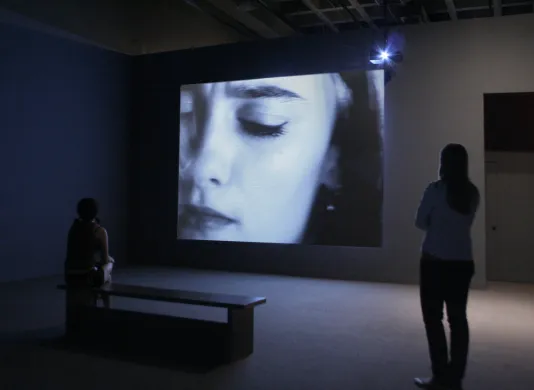2 people in a dark gallery watching a video installation with a close shot of a woman's face.