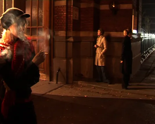 Video still of a woman smoking on a darkened street corner wtih 2 people on the far corner. 