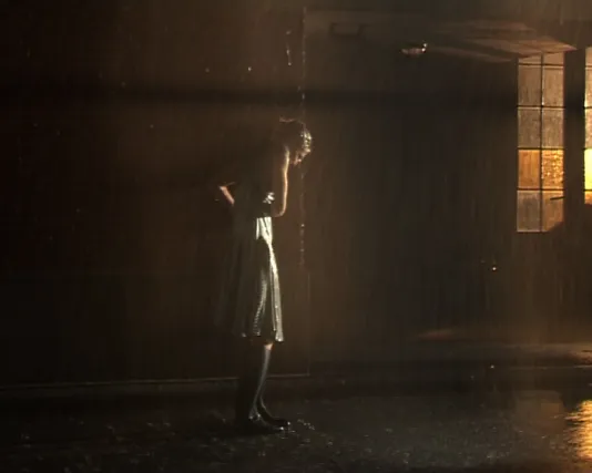 Video still of a woman standing outside in the pouring rain dressed in a light sun dress. 
