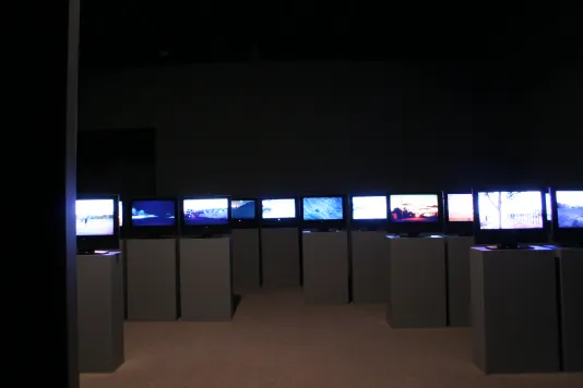 Many screens installed on podiums in a darkened gallery. 