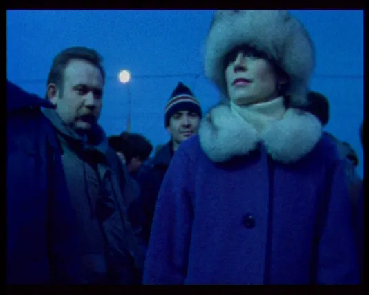 Video still of a woman in a fur hat with a matching blue coat in the foreground with 3 men standing right behind her.
