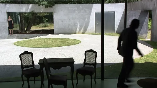 A man standing inside a modern home looking out to a courtyard with a woman in the distance. 