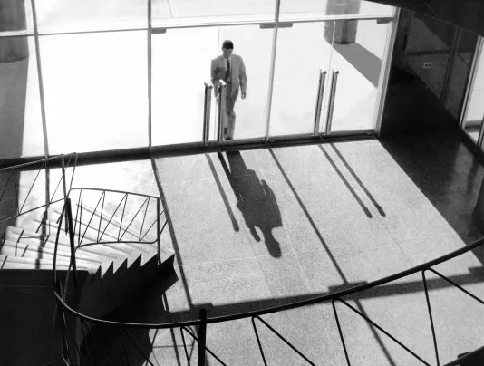 A black and white photo of a man in suit standing at tall glass doors seemingly trying to enter a building. 