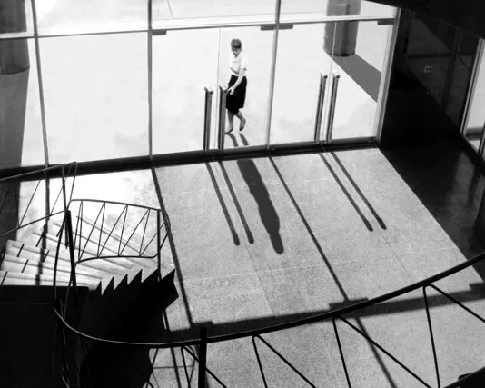 A black and white photo of a woman standing at tall glass doors seemingly trying to enter a building. 