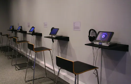 A wall of small TVs with individual headphones installed in a gallery.