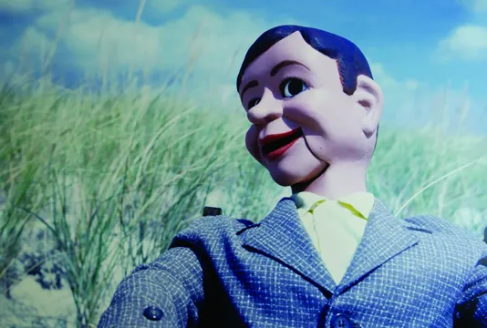 A close photograph of a ventriloquist dummy with wide eyes, smiling red lips, dressed in a suit jacket on a beach.