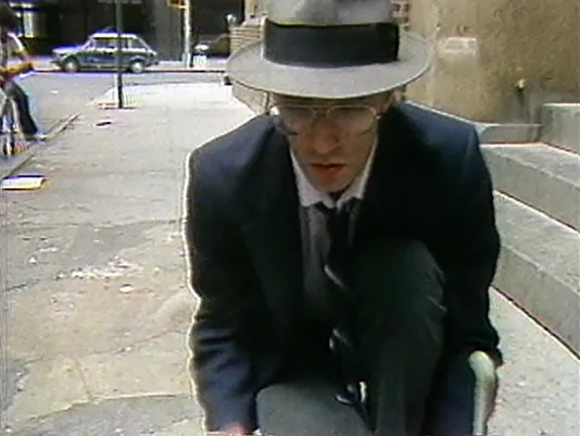 A video still shows a close view of a man dressed in a suit, tie, fedora hat and glasses kneeling on a city sidewalk. 