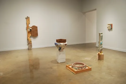 Five ceramic and wood sculptures; one hanging on a wall, one leaning against a wall, and three placed on the concrete floor