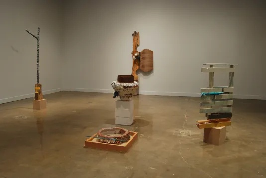 Five sculptures; three on low plinths, one leaning on a wall, and a flat, low sculpture with wood, a round object, and glass