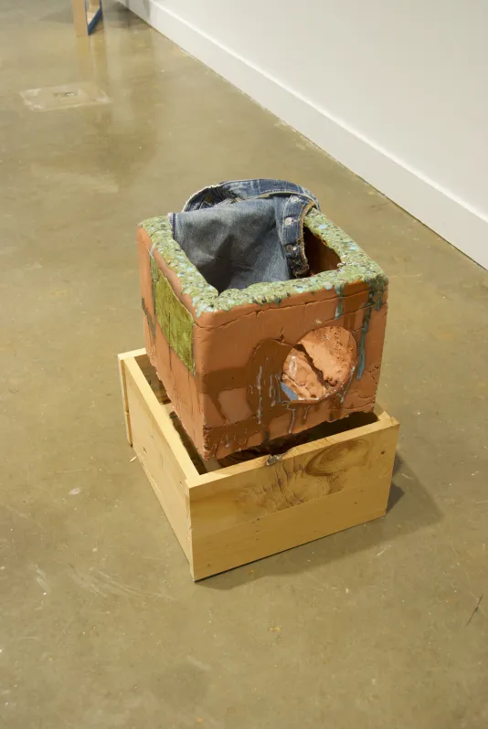 A floor sculpture with a square wood base, a smaller terra-cotta box on top, a piece of denim folded over at the very top.