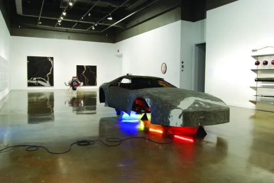 Installation view of a car shell without tires and lit from underneath with bright color tubes. Two paintings on back wall