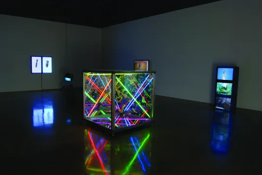 A darkened space with a large transparent cube structure lit from inside with bright colorful tubes, two video monitor sculptures