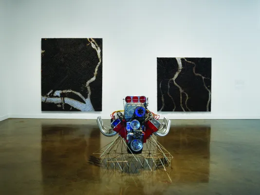 Two large mostly dark paintings of aerial views of city grids, and a large, ornate car engine on a base of thin metal rods