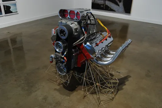 Close up of an ornate car engine sculpture with bright color wires, shiny pipes, and a stand of thin metal rods crisscrossed
