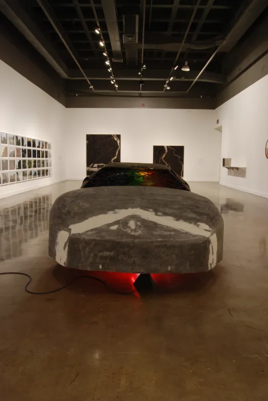 A long front view of a car shell with red light underneath, a grouping of pictures on a side wall, and two paintings in back