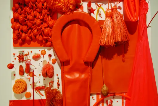 Close up of a white board covered with bright orange plastic objects; a horseshoe, tassels, fabric, a coil, and more