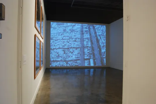 A darkened space with an aerial image of a city projected on the front wall, and framed pictures on the adjacent wall