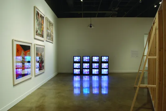 Twelve stacked video monitors with the same image, the back of a wood framework, and four framed photos on the wall